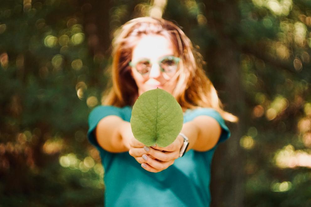 A woman in a teal top holding a large green leaf directly in front of her, with her face obscured by the leaf and a blurred natural background, highlighting a connection with nature.