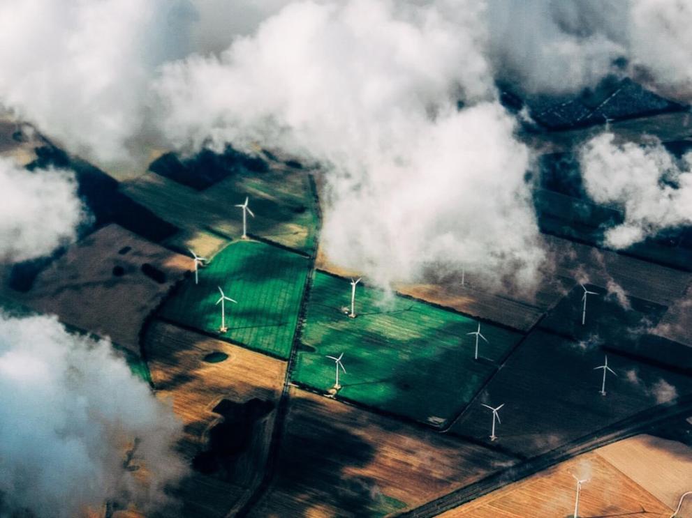Aerial view of a wind farm with turbines dotted among a patchwork of green and brown fields, partially veiled by white clouds, illustrating the integration of sustainable energy production with rural farming.