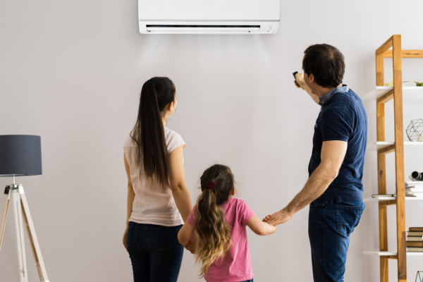 A family inside a home with a modern decor, standing together and looking at an air conditioning unit on the wall, with the father adjusting the settings with a remote control