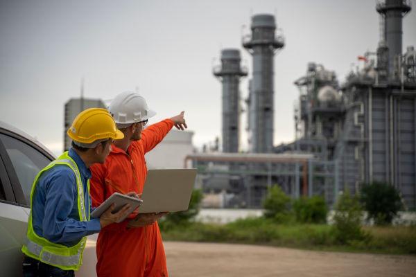 Two construction workers are standing in front of a large industrial plant.