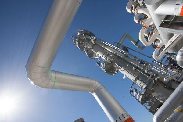 The image shows a close-up of a large industrial pipe with other smaller pipes branching off in different directions with what look like a chemical plant or refinery in the background over a clear light blue sky with a bright sun shining from the left side.