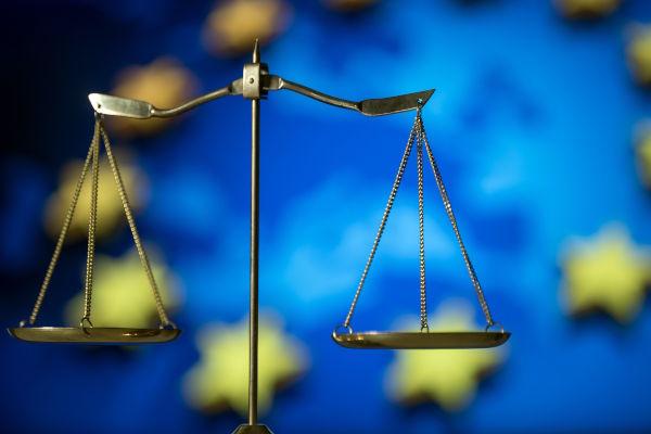 The image shows a close-up of a balanced scale in front of a blurred European Union flag suggesting legal proceedings or legislation. The image evokes a sense of justice, balance, and fairness.